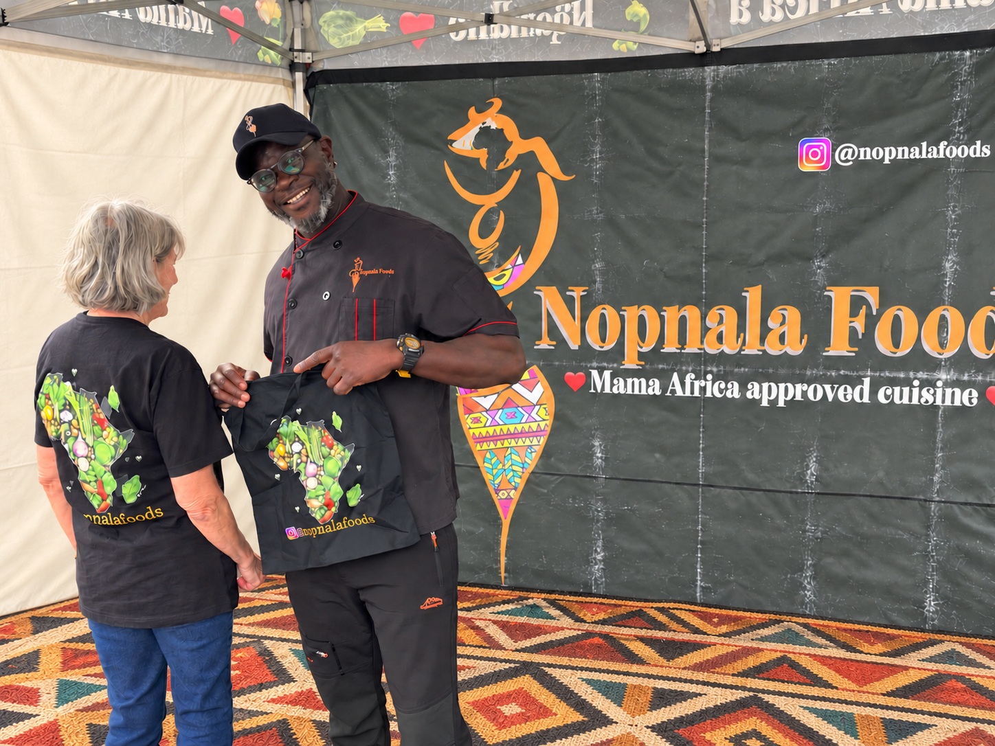Hajj at the Nopnala Foods market stall beside Cynthia wearing the Africa and vegetable design on her shirt, while he holds a matching cloth bag