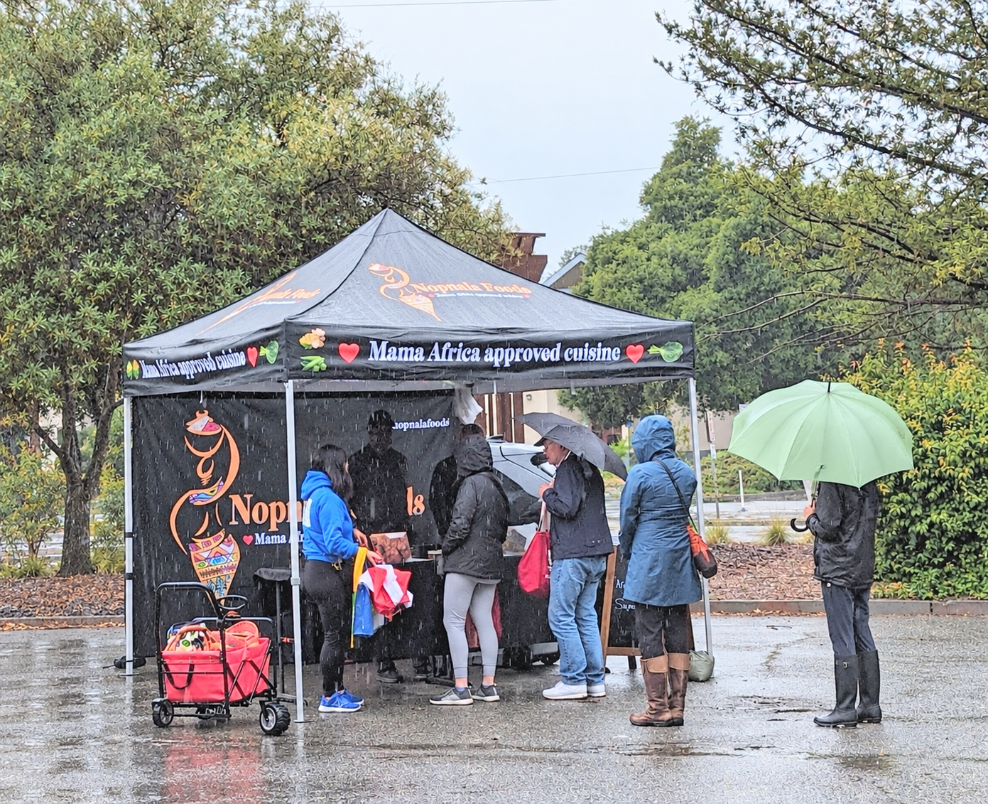 Customers waiting with umbrellas at the Nopnala Foods tent on a rainy market day
